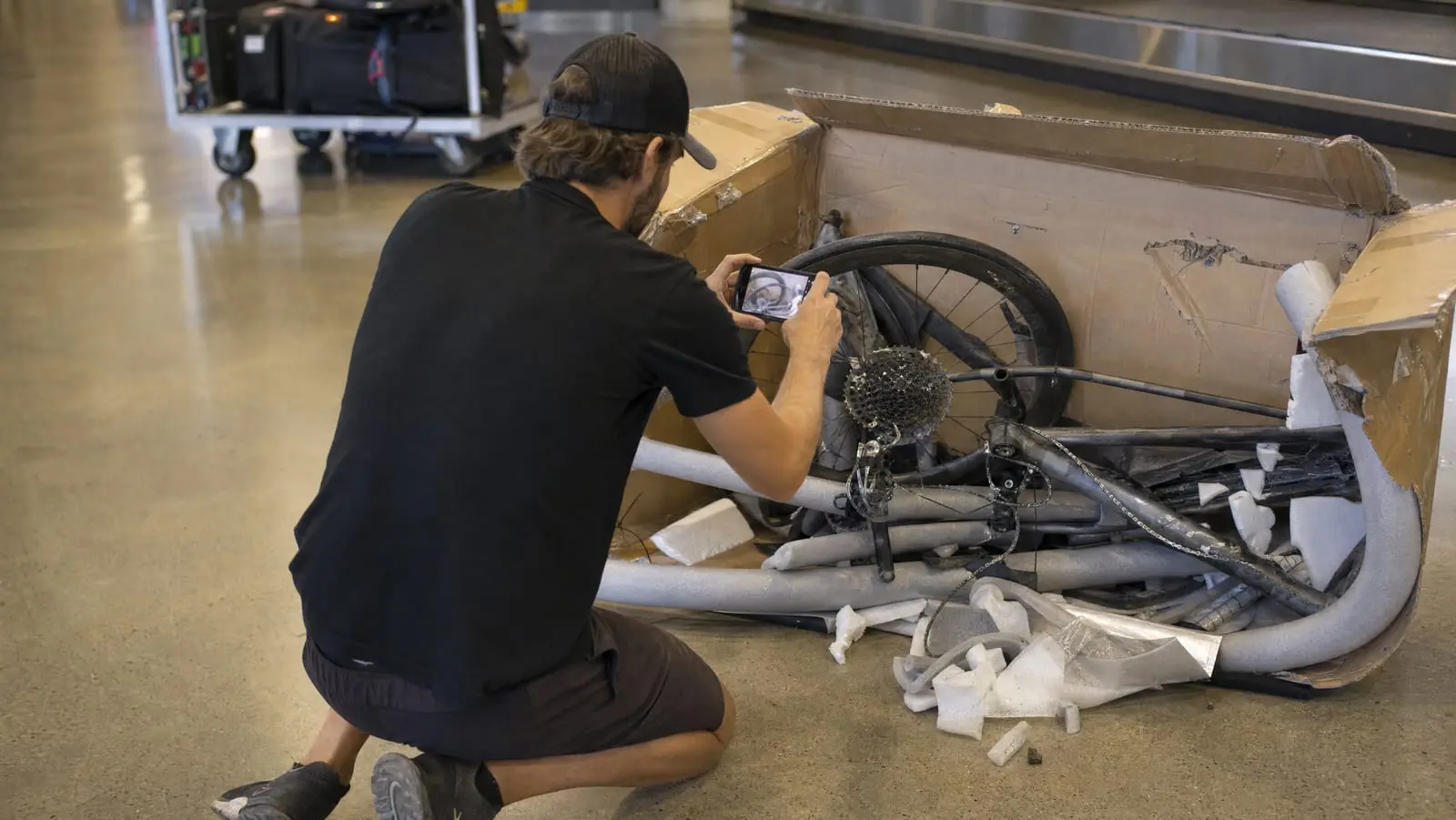 A cyclist photographing a bent rear derailleur beside an opened cardboard bike box in an airport baggage area