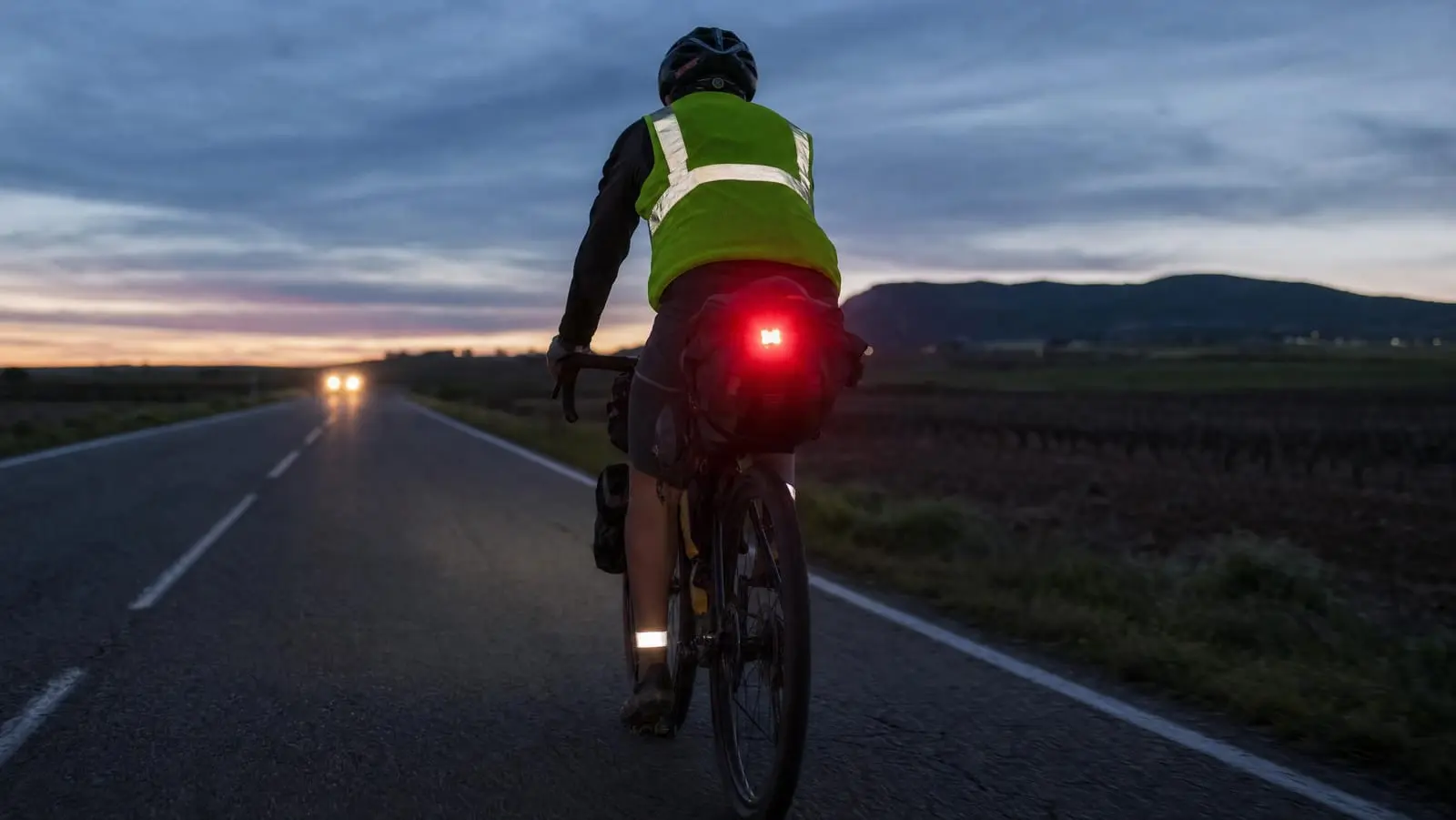 Loaded bikepacking rider seen from behind on a rural road at dusk, rear light glowing and reflective ankle bands and vest flaring in an approaching cars headlights