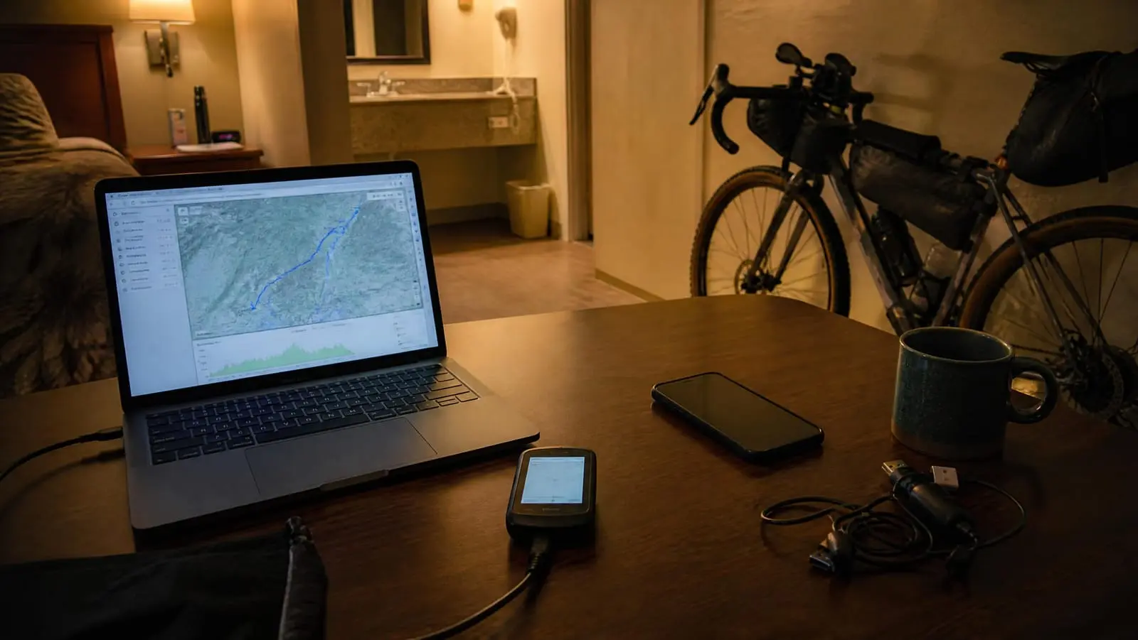 A motel desk at night with a bike computer plugged by USB into a laptop showing a route map, a phone and charging cables beside it, a loaded bike against the wall