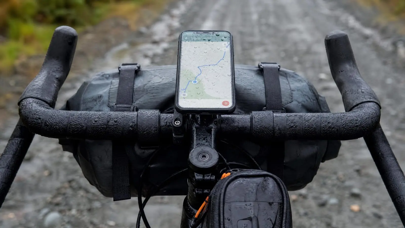 Close view of a loaded bikepacking cockpit with a smartphone in an out-front handlebar mount showing a navigation map, rain beaded on the screen above a handlebar roll